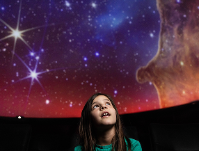 A young girl looking up at a colorful starry sky in a planetarium.
