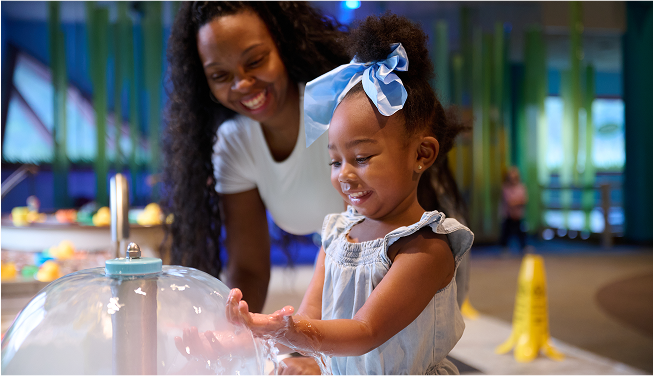 A mother smiles watching her young daughter experiment with a water exhibit.