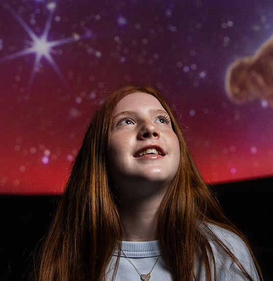 A young girl looking up at a colorful starry sky in a planetarium.