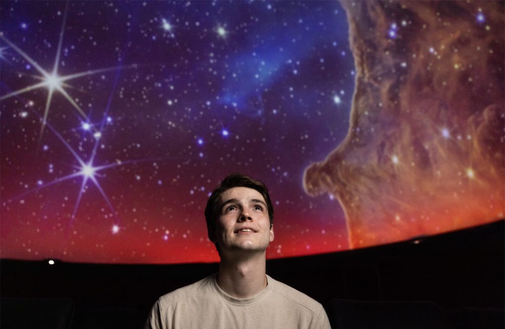 A young man looking up at a colorful starry sky in a planetarium.